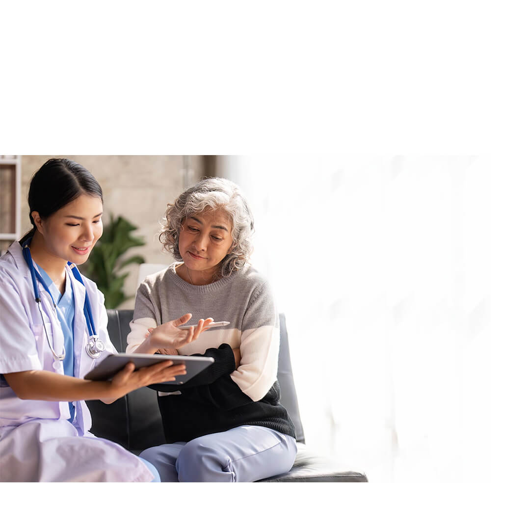 A nurse and an older adult patient sit together reviewing information on a tablet in a consultation room