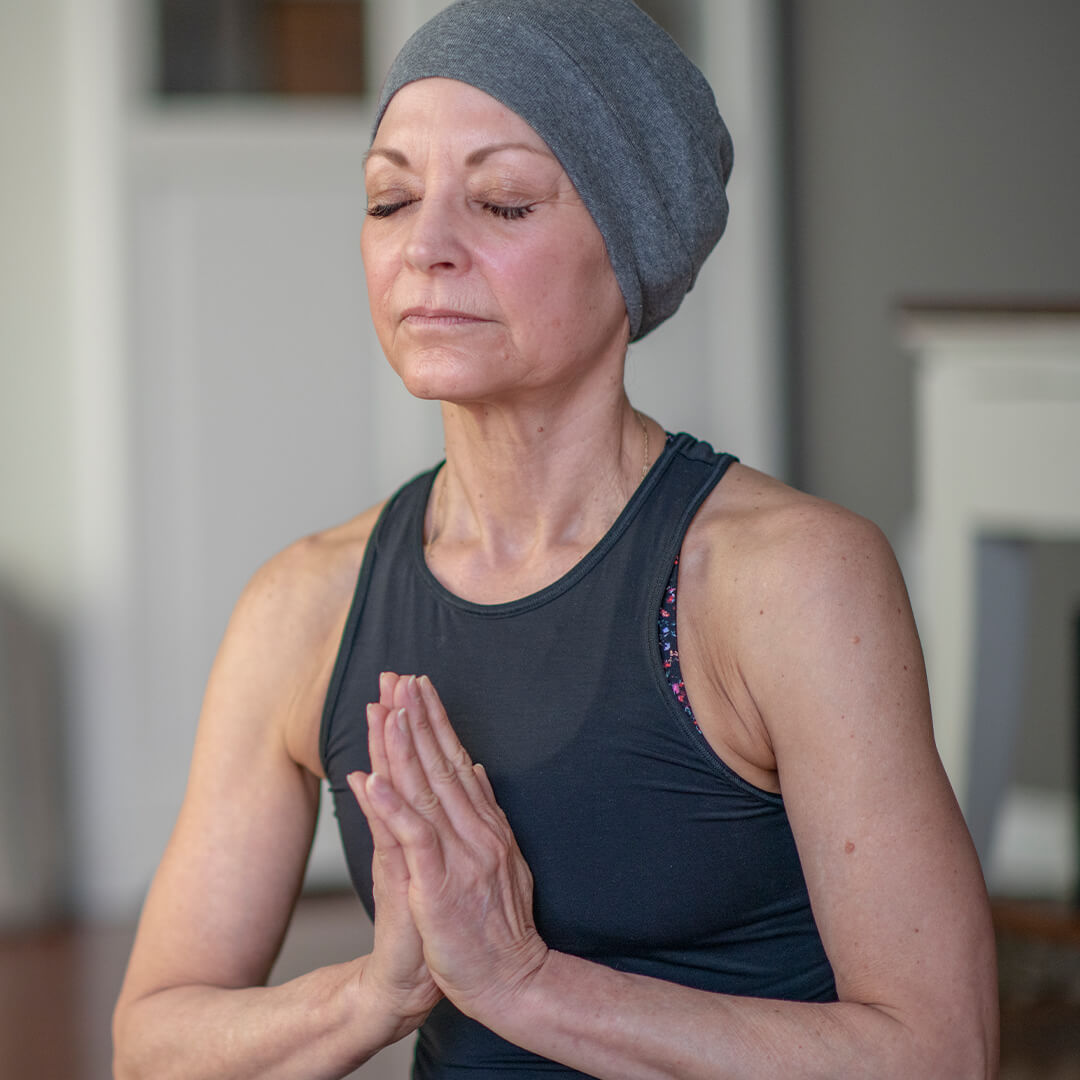 Middle-aged woman wearing a headscarf sits cross-legged with closed eyes and hands pressed in prayer, practicing mindfulness for cancer wellness