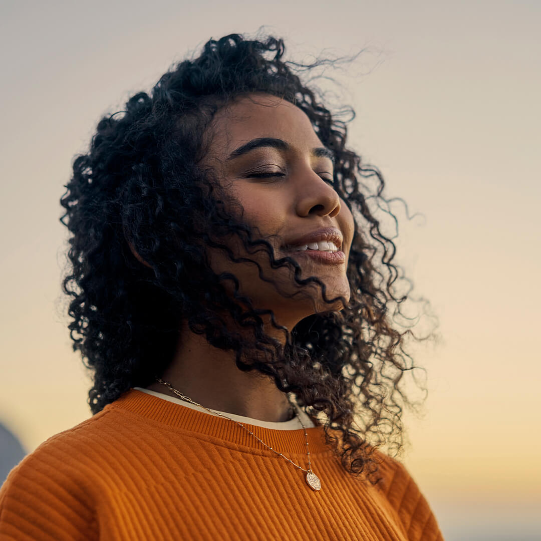 Young woman closes her eyes, reflecting peacefully outdoors at sunset, embodying spiritual strength in a cancer journey