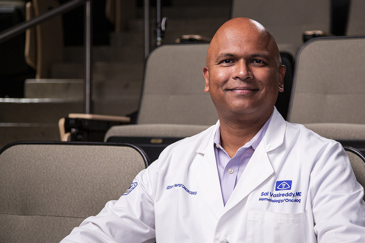 Dr. Sal Vasireddy, a smiling oncologist in a white coat, sits in a lecture hall setting conveying warmth and professionalism